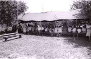 A black and white photo of people under the canopy, eating lunch.  There are benches in the foreground, where the business meeting will be held.  Date for this photo is unknown.  Thanks to Anne Follweiler.